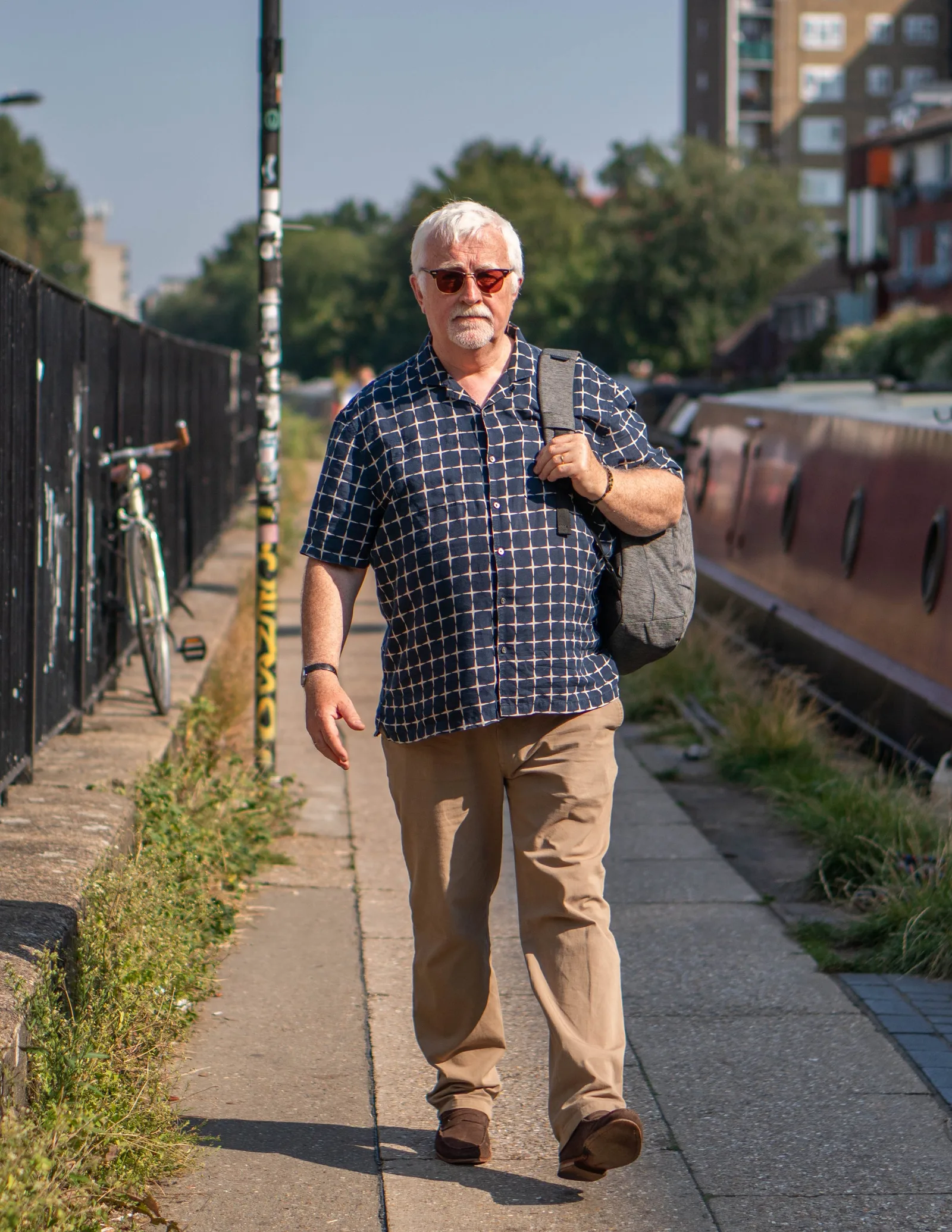 A white-haired man in sunglasses and a checked shirt walking along a canal path, a bag over his shoulder