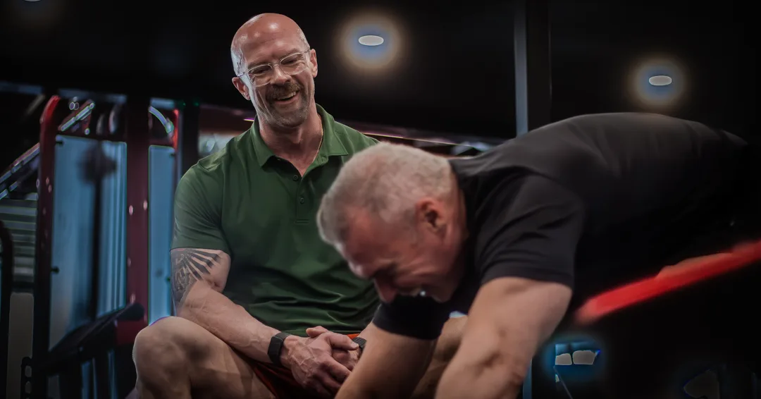Tyson DesMarais seated in his gym, talking with a client who is working beside him.