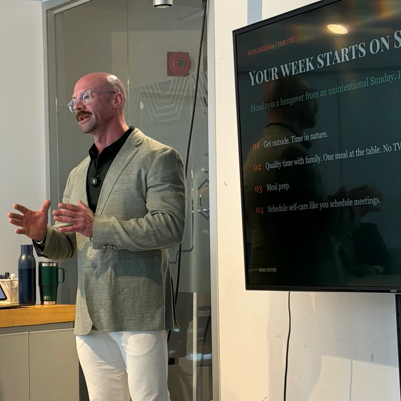 Tyson DesMarais presenting to a workplace room, head up and mid-sentence, hands open, his own slide on the screen beside him